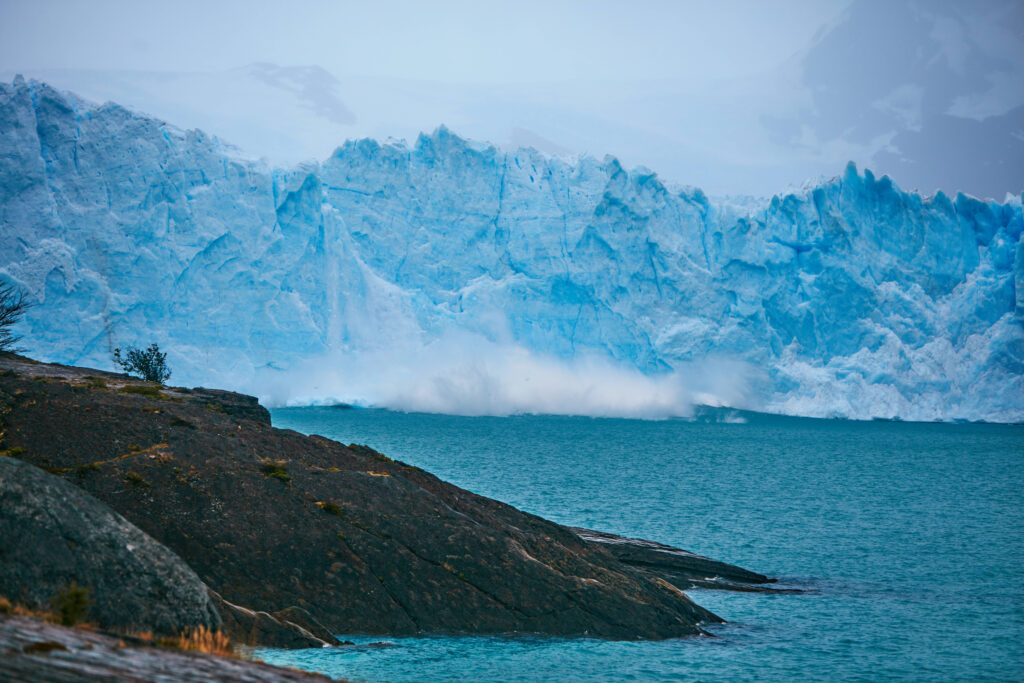 Massive blue iceberg towering over turquoise Arctic sea with rugged dark rocks in foreground Wallpaper