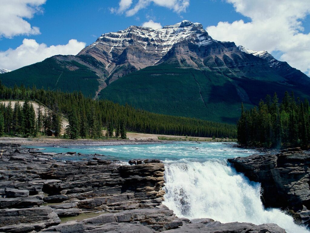 Majestic Canadian waterfall cascades over rocks with evergreen trees and snow-capped mountains Wallpaper