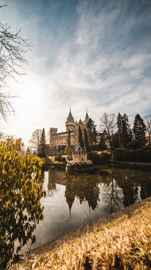 Majestic castle with towering spires reflected in lake surrounded by greenery and winter trees Wallpaper