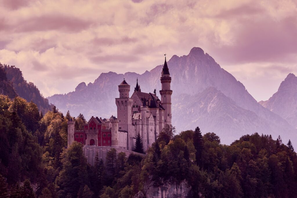 Majestic castle with elegant towers above lush forest and rugged mountains in soft pink hue wallpaper