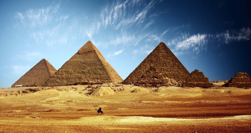 Giza Pyramids under vibrant blue sky with wispy clouds and lone rider in golden desert landscape Wallpaper