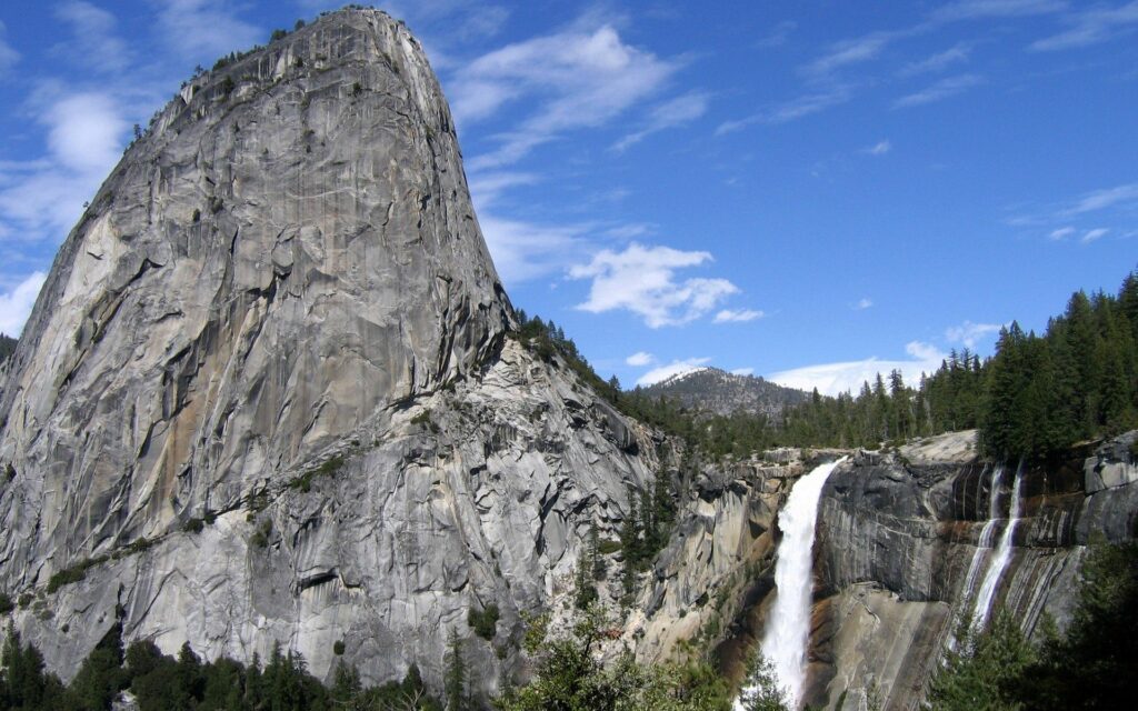 Massive gray rock formation and waterfall surrounded by giant stones and lush green trees Wallpaper