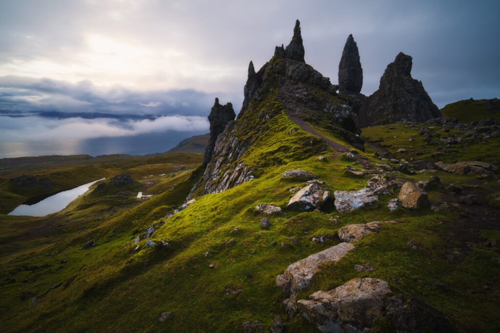 Jagged rocks on grassy slope overlooking serene valley with sunlight on green terrain and water Wallpaper