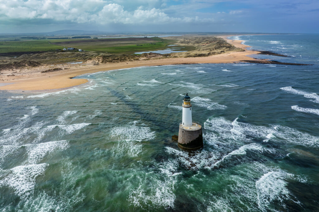 Majestic lighthouse on rugged coast with foamy waves sandy beaches and green fields under sky Wallpaper