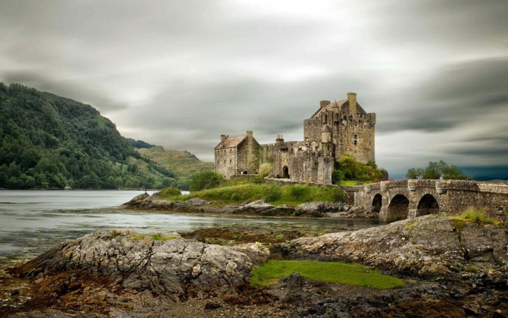Majestic medieval castle by water at sunset surrounded by greenery and rugged rocks wallpaper
