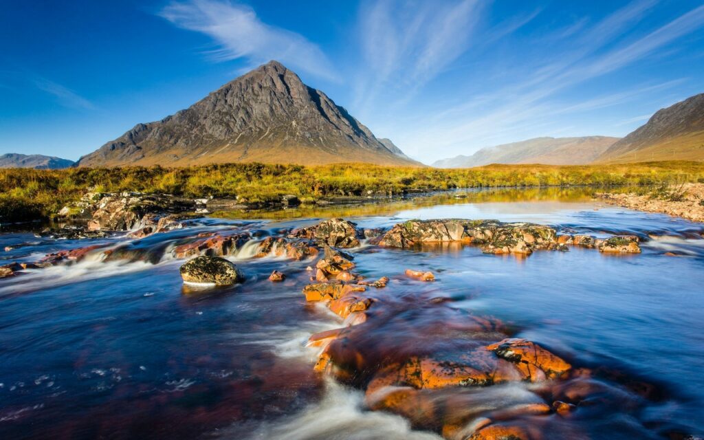 Majestic mountain landscape with clear river and vibrant rocks under vivid blue sky and clouds Wallpaper