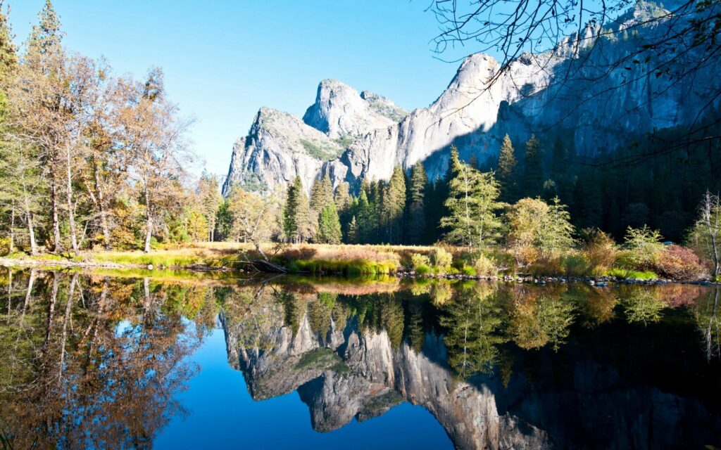 Majestic mountains reflected in a tranquil lake with lush forests under a clear blue sky wallpaper