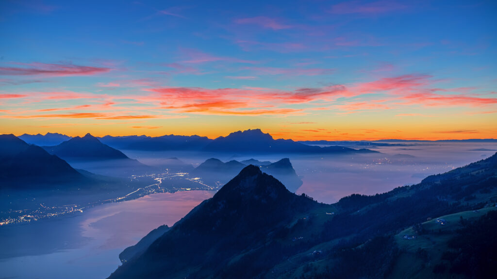 Majestic mountain peaks at sunset over misty lake with glowing riverside town landscape wallpaper