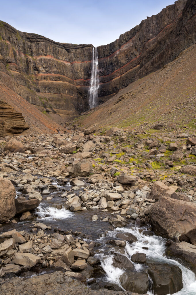 Tall narrow waterfall cascading over rugged rocks into clear stream surrounded by stones and earth Wallpaper