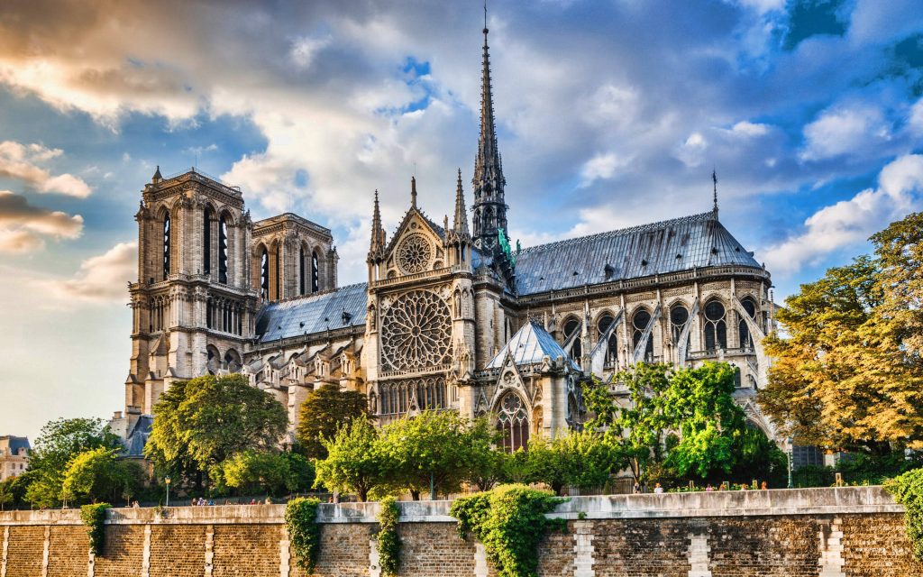 Notre-Dame de Paris Gothic cathedral with rose window and spire lit by sunlight by riverside trees Wallpaper