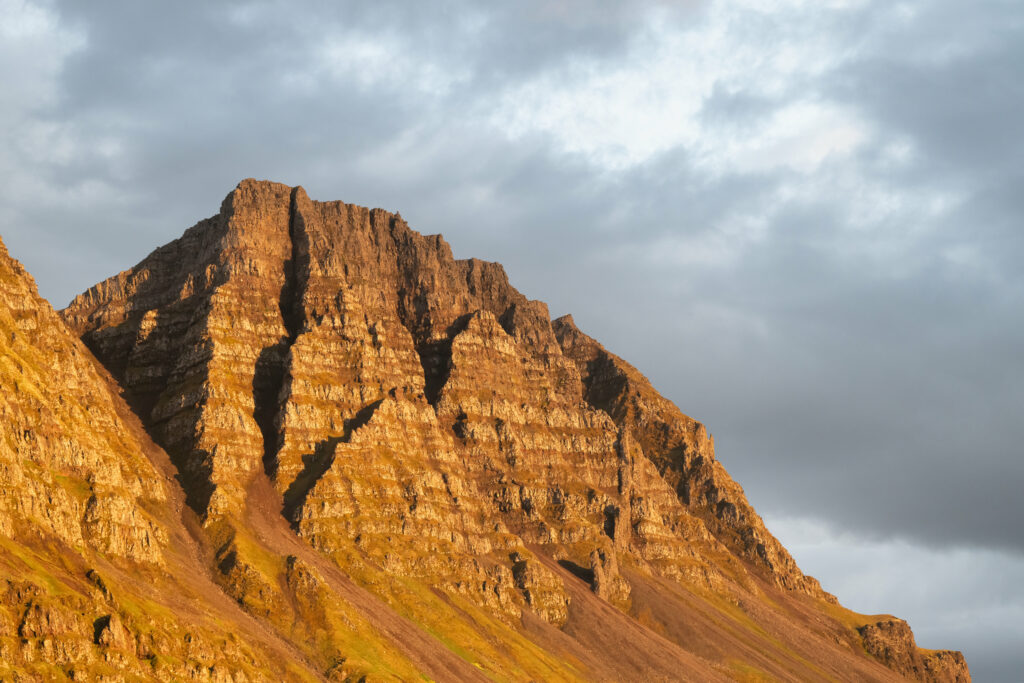 Rugged mountain peak glowing in warm evening light with textured rocks and moody clouds background Wallpaper