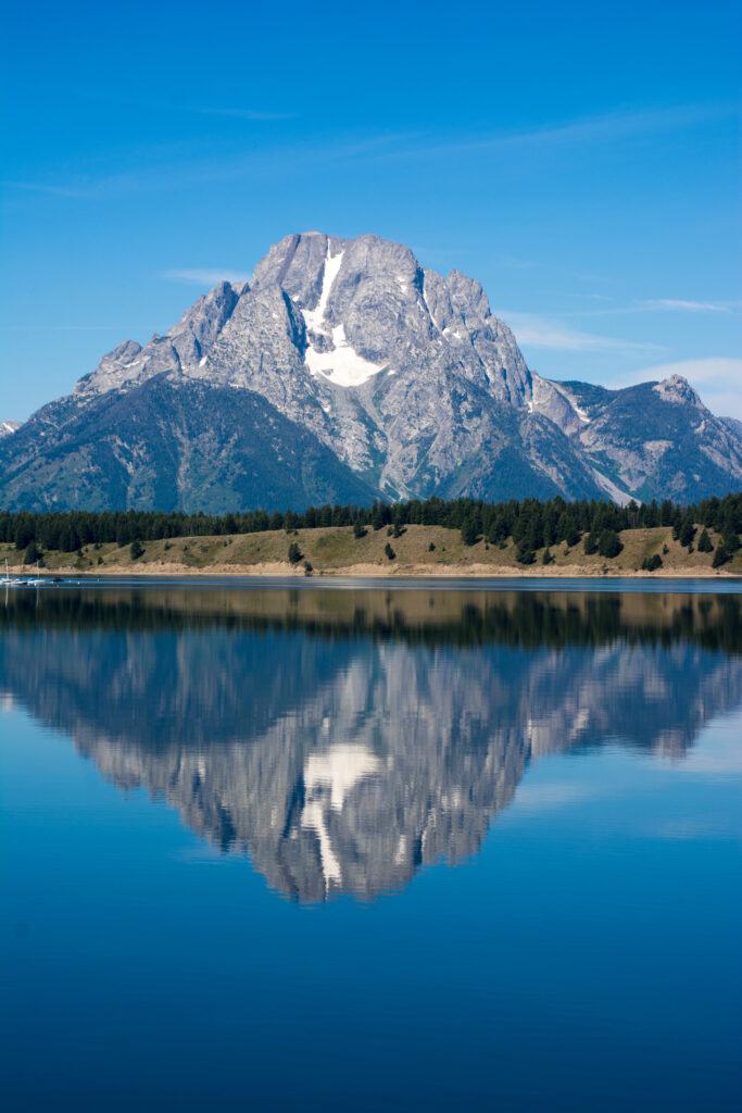 Snow-capped mountain reflected in clear lake with lush forest and vibrant blue sky background Wallpaper