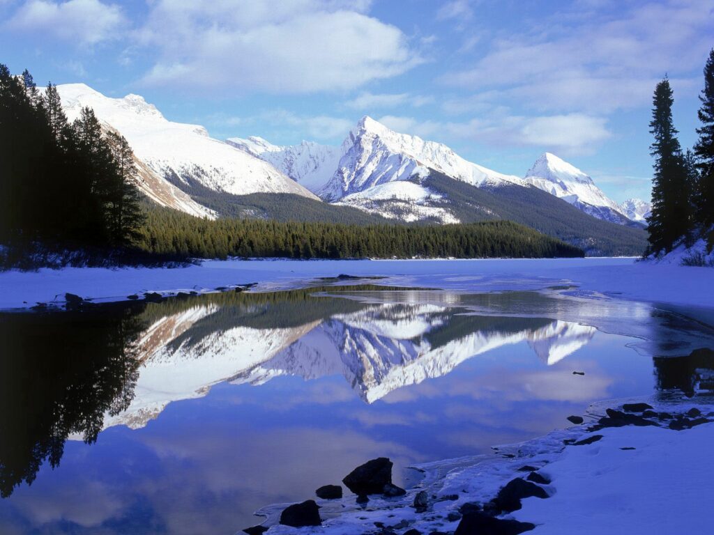 Snow-capped mountains reflected in a still lake with evergreen forests under blue sky wallpaper