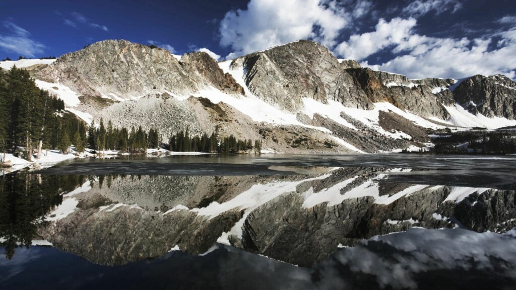 Snow-capped mountains reflected in calm lake with blue sky and evergreen trees wallpaper background