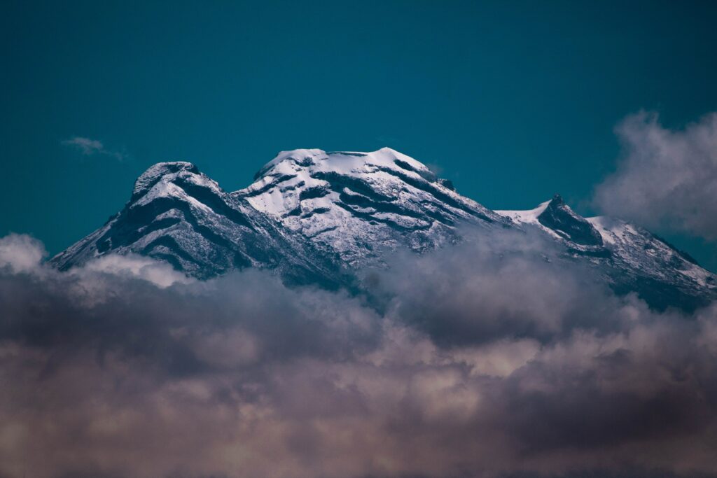 Snow-capped volcanic mountain peak above clouds under deep blue sky with dramatic lighting contrasts Wallpaper