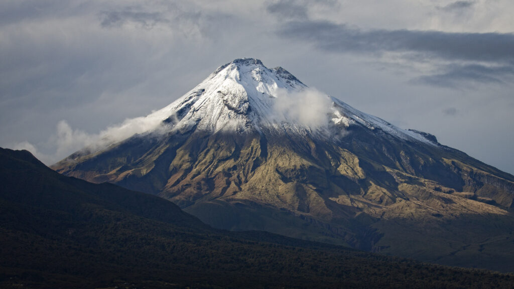 Snow-capped volcano above rugged mountains with drifting clouds in serene natural landscape Wallpaper