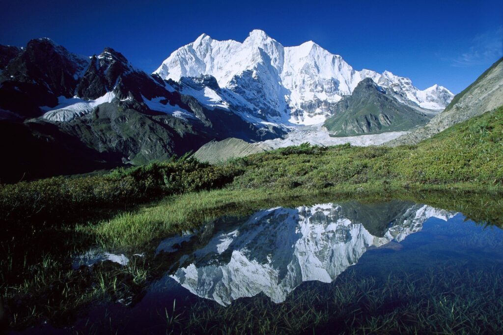 Snow-capped Asian mountains above green fields with clear blue sky and reflective water wallpaper