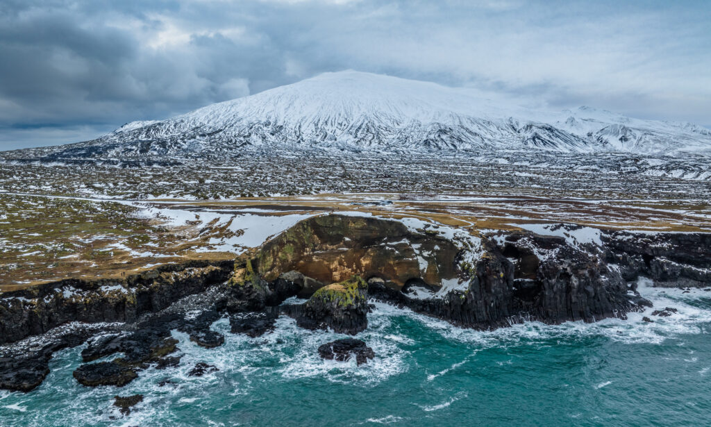 Snow-capped mountain above rugged rocky coast with icy waves crashing in serene natural landscape Wallpaper