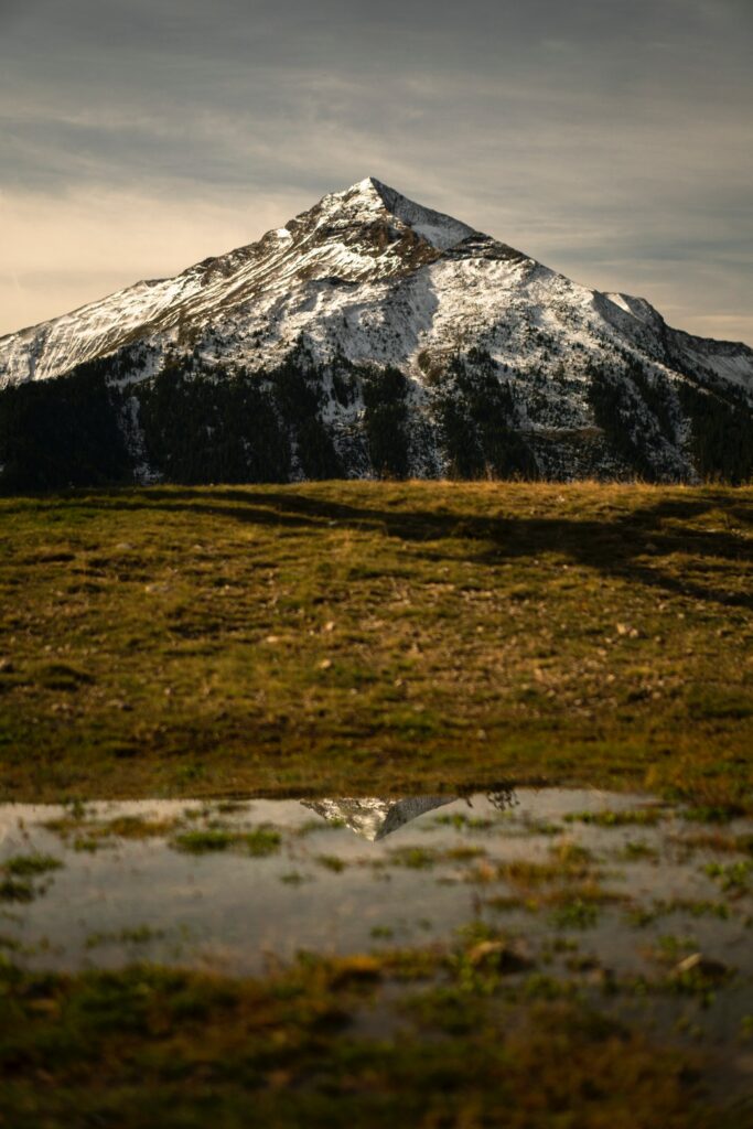 Snow-capped mountain peak above green field reflected in clear puddle serene nature wallpaper background