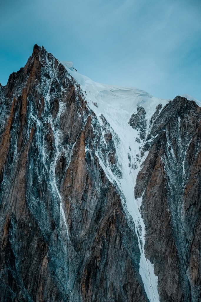 Rugged snow-dusted mountain peak with steep rocky cliffs against a clear blue sky wallpaper background