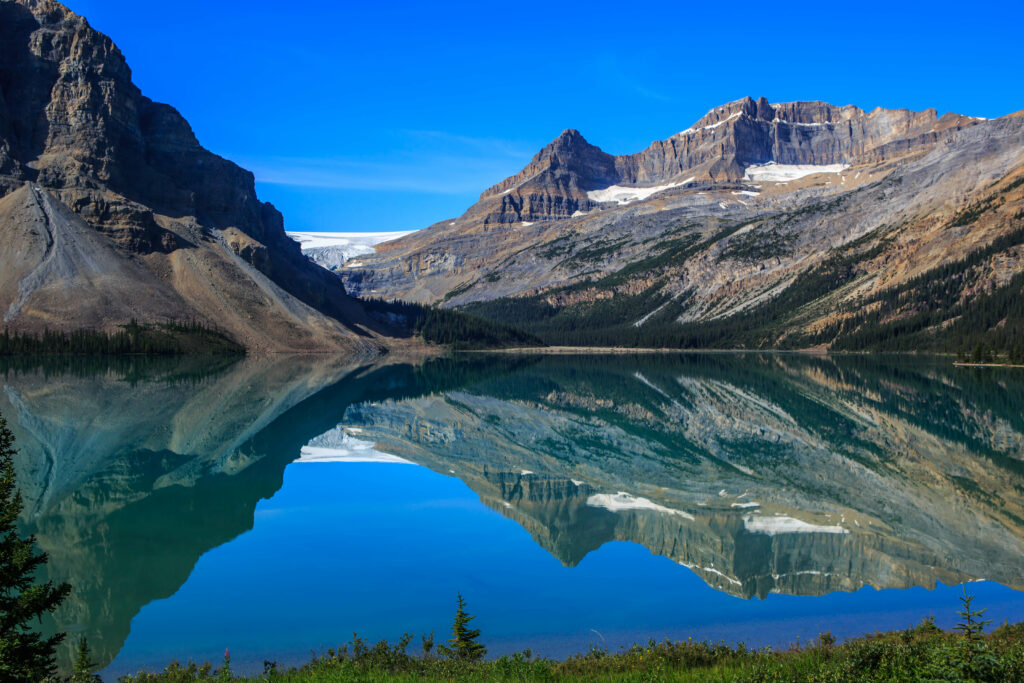 Snow-capped mountains reflected in crystal-clear lake with evergreen trees under blue sky wallpaper