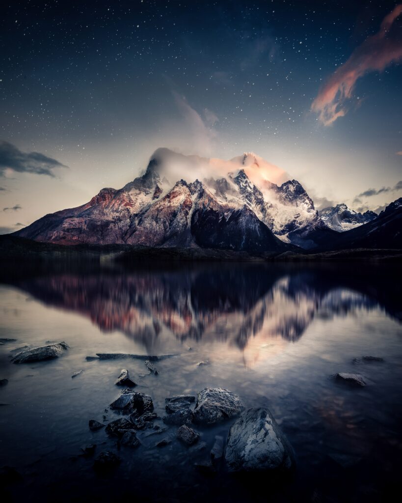 Snow-capped mountains under starry sky reflected in calm lake with smooth stones foreground wallpaper