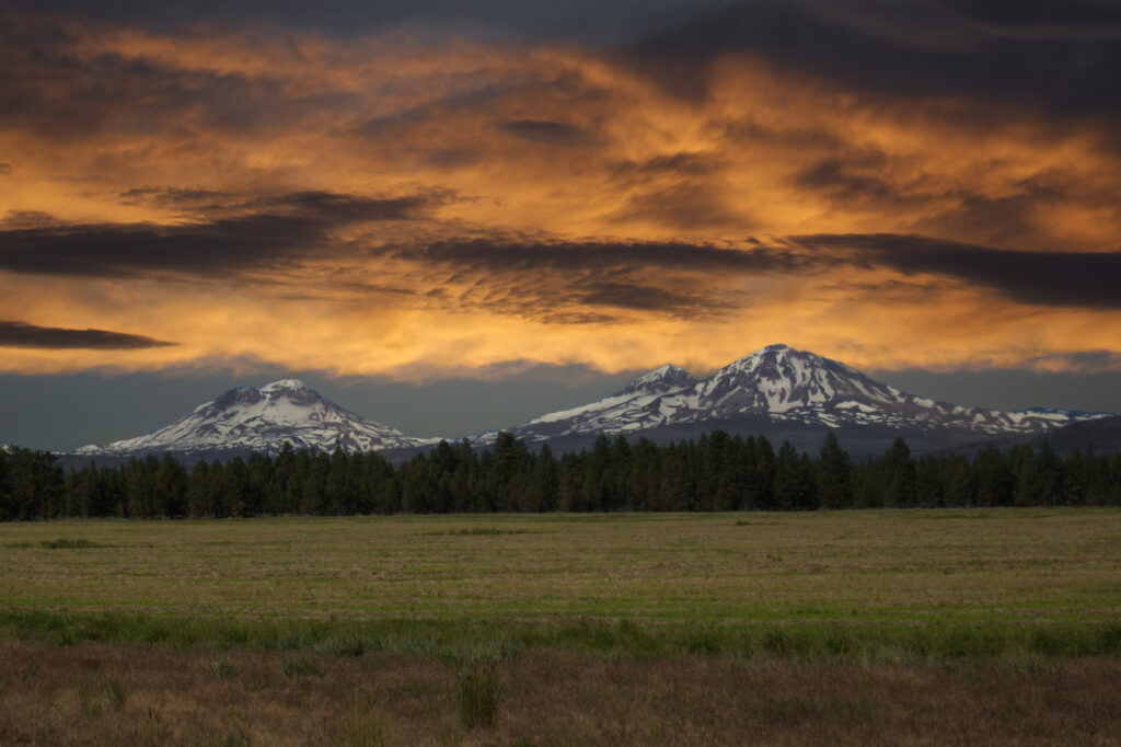 Snow-capped mountains with rugged rocks above forest and field under dramatic orange sky wallpaper