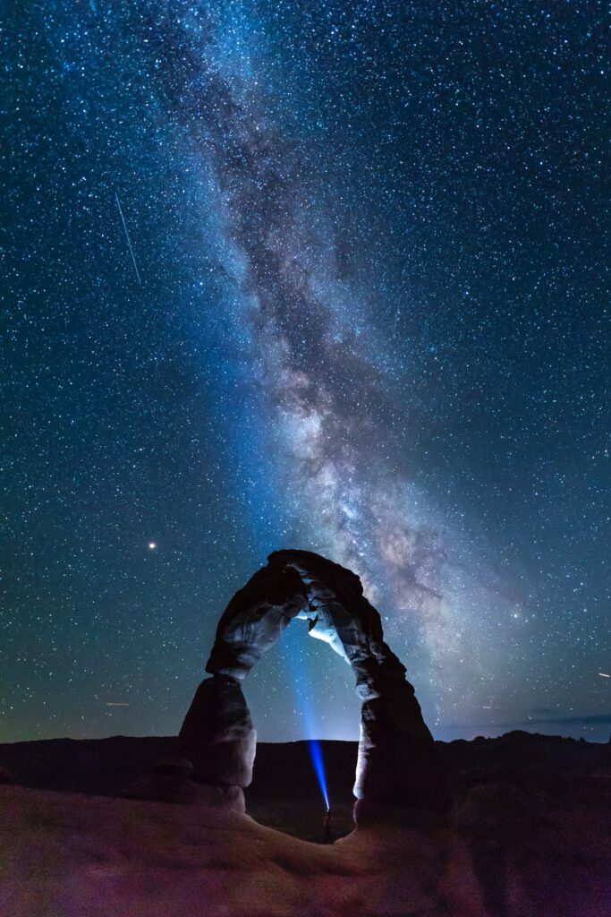 Majestic stone arch at night with starry sky and distant figure’s flashlight illuminating rocks Wallpaper
