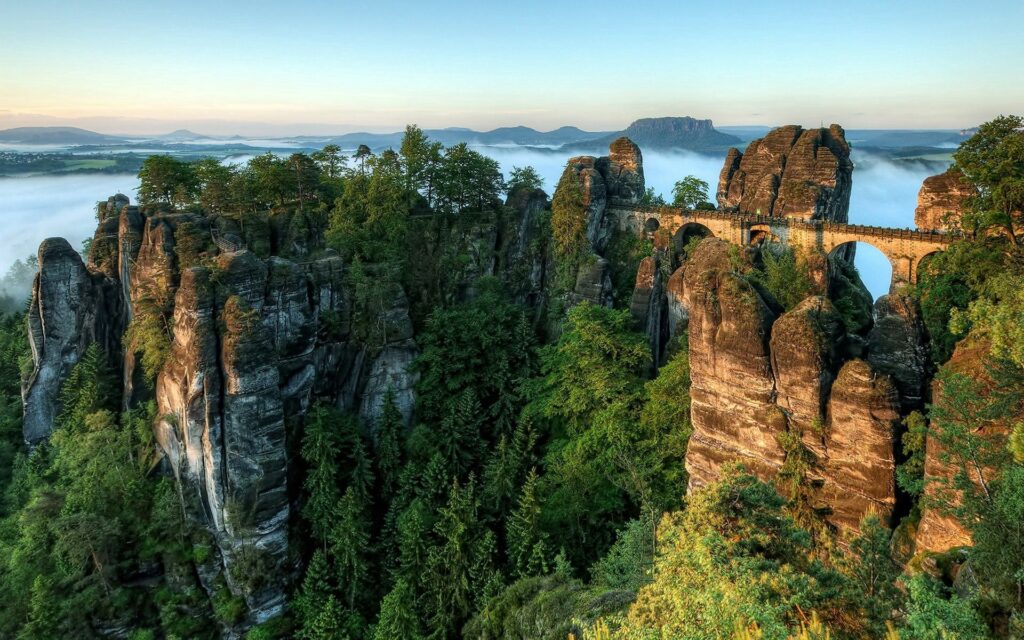 Majestic stone bridge over towering rocks with lush pine forests and misty clouds background Wallpaper