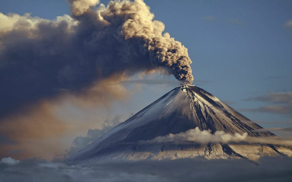 Majestic volcano eruption with dark smoke plume over snowy rugged slopes under clear sky Wallpaper