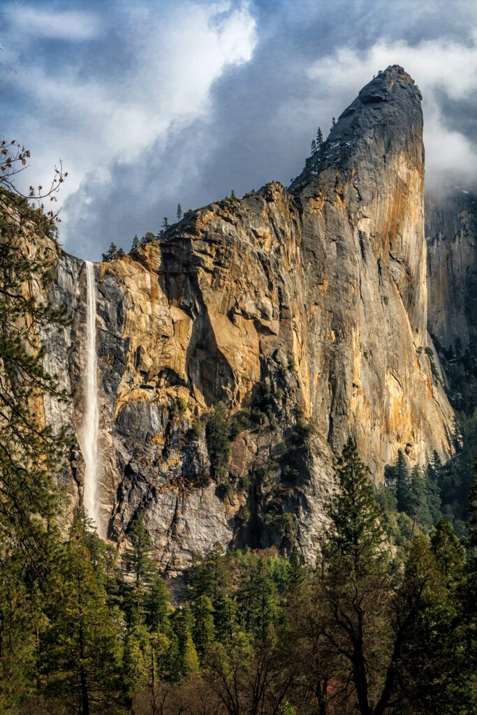 Majestic waterfall cascading down cliffs framed by lush green trees under dramatic cloudy sky Wallpaper