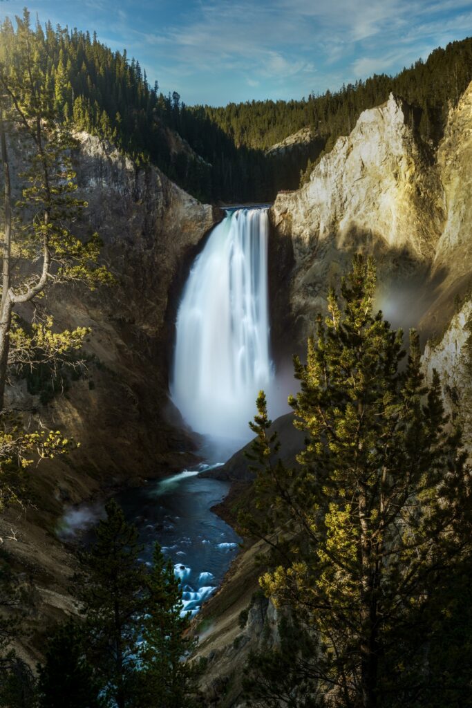 Majestic waterfall cascading down rocky cliff with sunlight mist and lush green pine forest background Wallpaper