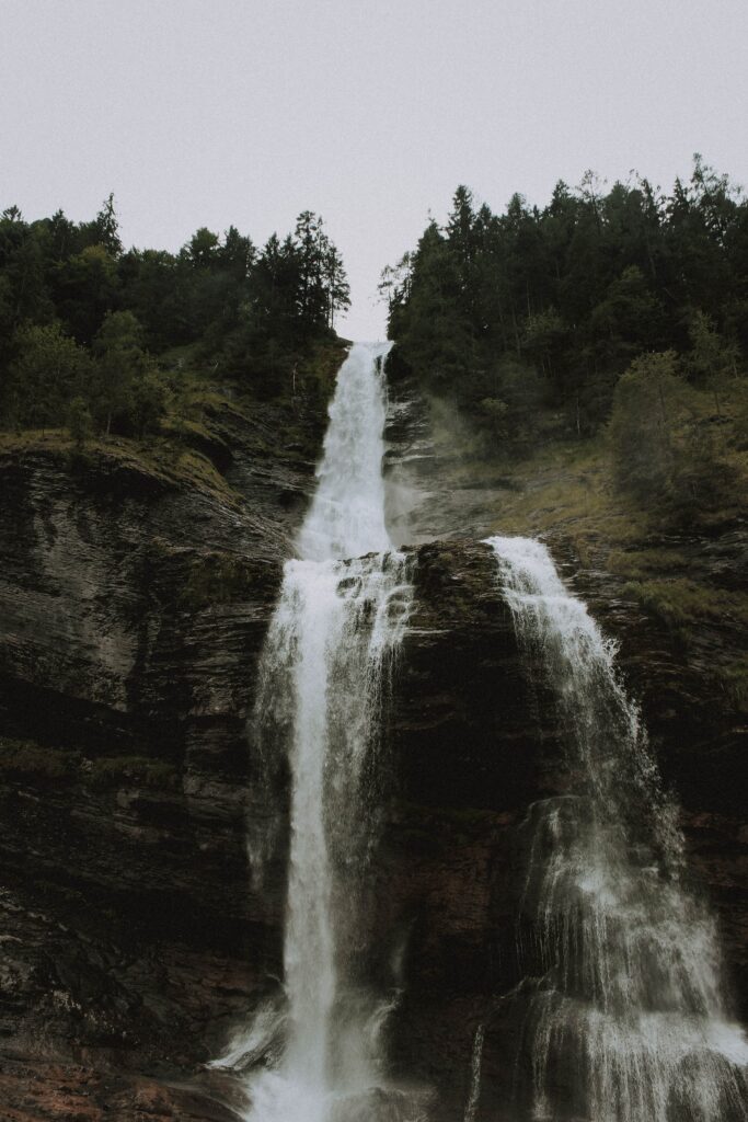 Majestic waterfall cascading down rocky cliff surrounded by lush green trees and rising mist Wallpaper