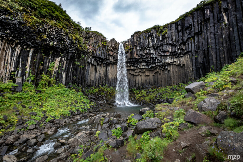 Tall waterfall cascading from basalt columns into clear stream surrounded by lush green landscape Wallpaper