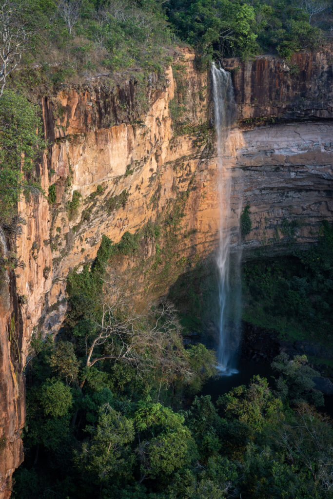 Tall waterfall cascading orange cliffs into green forest sunlight highlights textured rocks Wallpaper