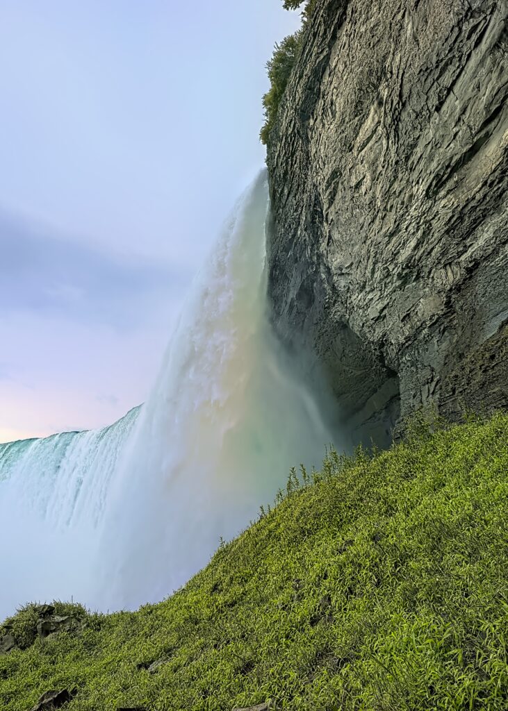 Waterfall cascading over rocky slope with vibrant green grass and pale blue sky background Wallpaper