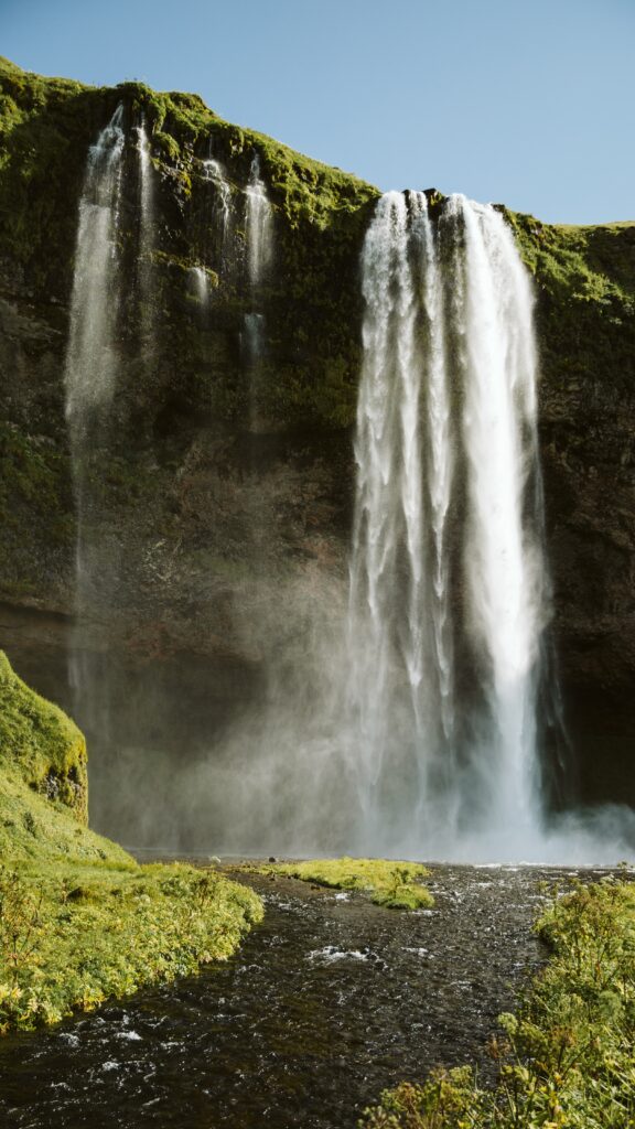 Majestic waterfall cascading rugged cliff surrounded by lush greenery under clear blue sky wallpaper