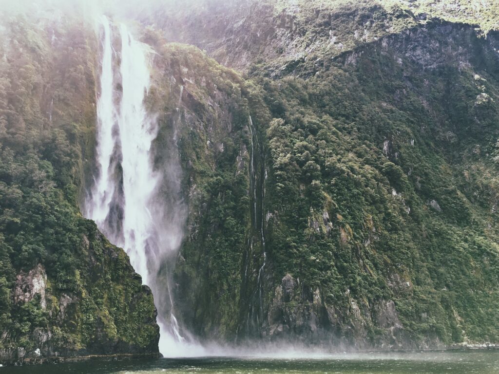 Majestic waterfall cascading down rugged cliff with lush green mountains and misty river below Wallpaper