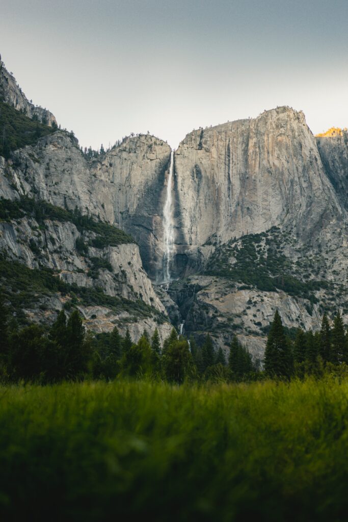 Majestic waterfall cascading rugged cliffs with lush forest and grass under soft natural light Wallpaper