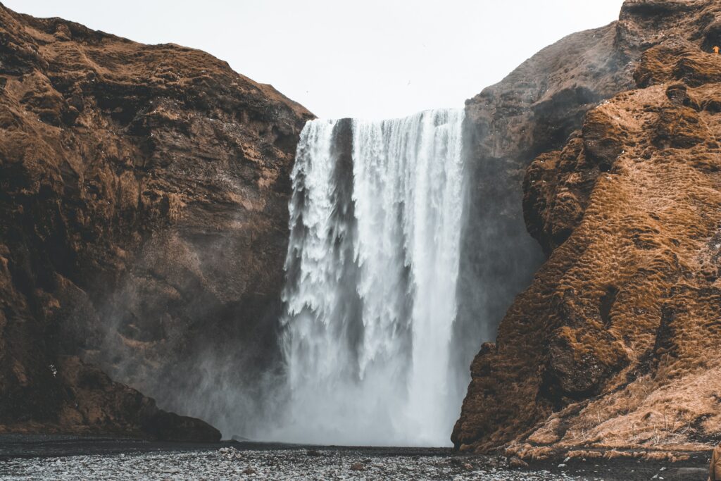 Majestic waterfall cascading over rugged brown cliffs with misty spray and rushing river below Wallpaper