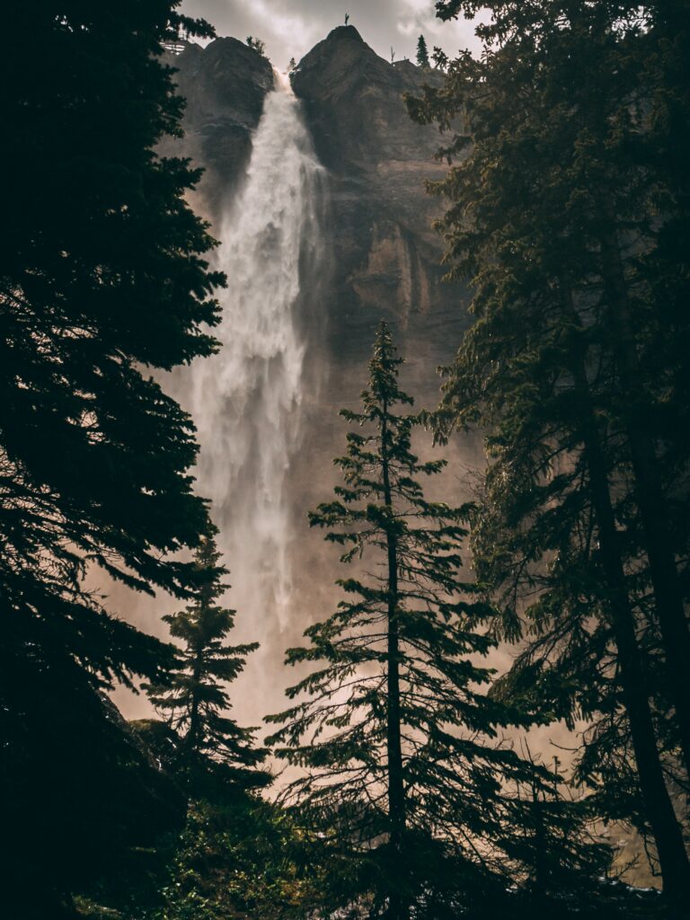 Majestic waterfall flowing down rocky cliff surrounded by lush green trees and soft sunlight mist Wallpaper