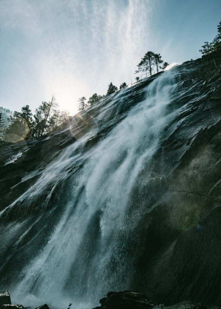 Majestic waterfall cascading down rugged cliff with lush forest and sunlight lens flare effect Wallpaper