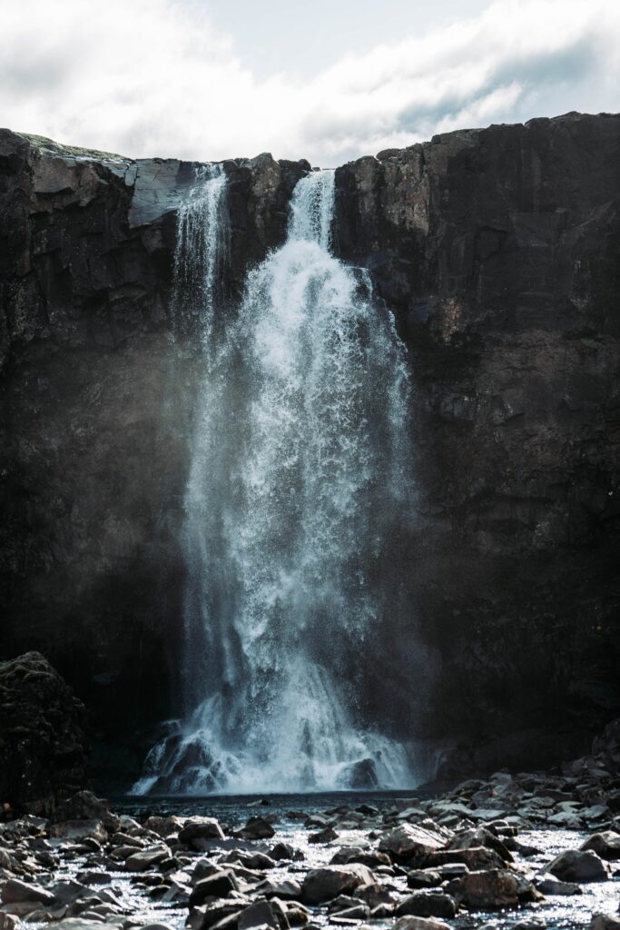 Powerful waterfall cascading over rugged cliff with clear water and dark stones under cloudy sky Wallpaper