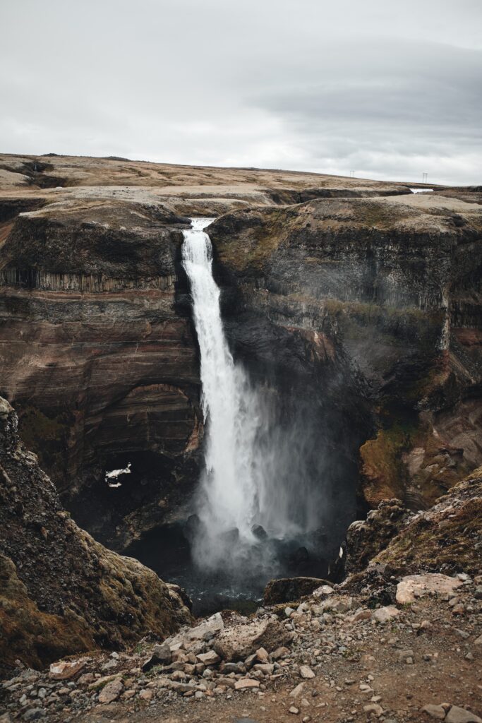 Waterfall cascading from rocky cliff into dark basin with natural earthy tones and textures Wallpaper