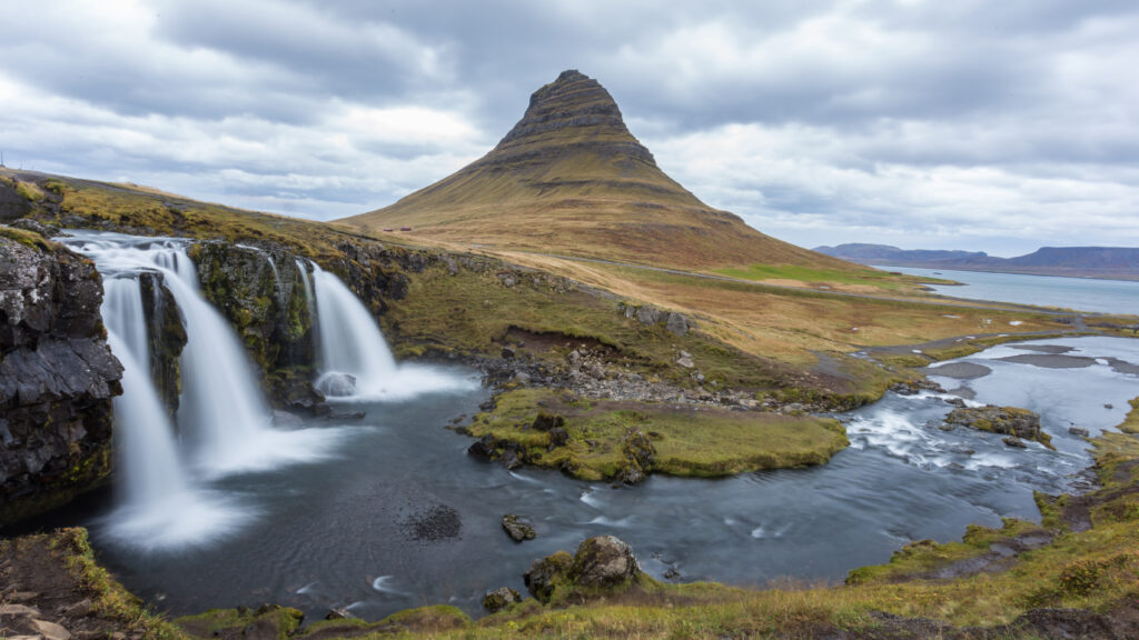 Cascading waterfall over rugged rocks into river with mountain peak and cloudy sky wallpaper