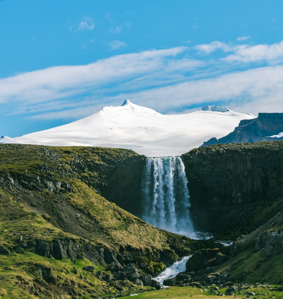 Majestic waterfall over dark rocks into green valley with snowy mountains and bright blue sky background Wallpaper