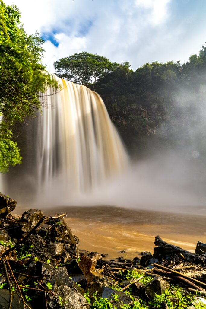 Powerful waterfall over rocky edge surrounded by lush green trees and sunlit mist landscape Wallpaper