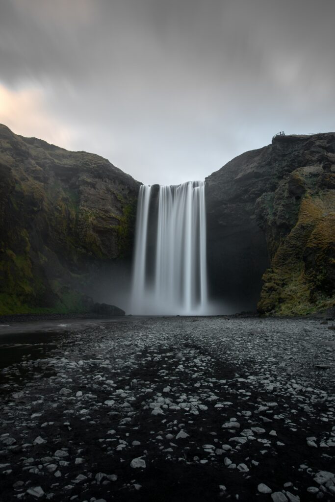 Majestic waterfall cascading over rugged cliff into serene river with mist and pebbled shore Wallpaper