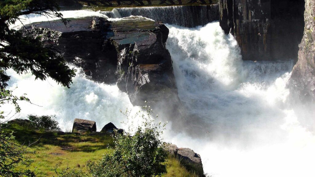 Powerful waterfall rushing over rocks with foamy water lush green trees and sunlight spray wallpaper