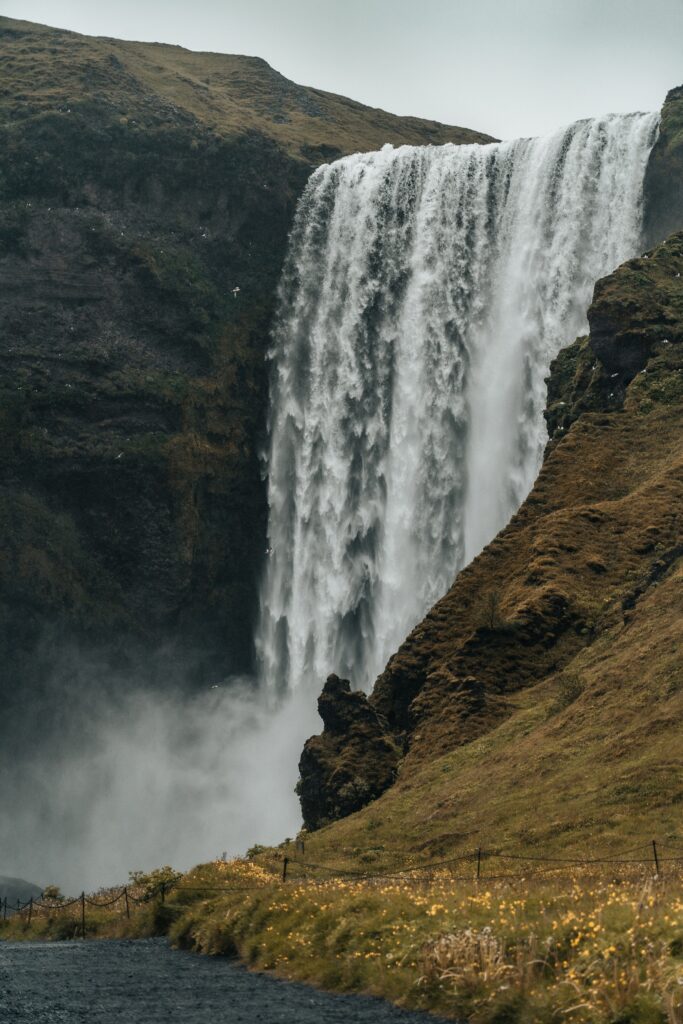 Majestic waterfall cascading over rugged rocks with mist surrounded by green grass and wildflowers Wallpaper